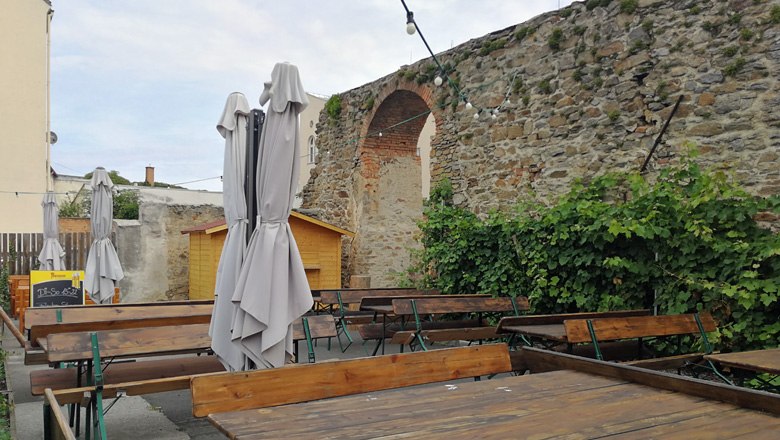 An empty outdoor area of a wine tavern with wooden tables, closed umbrellas and an old stone wall in the background.