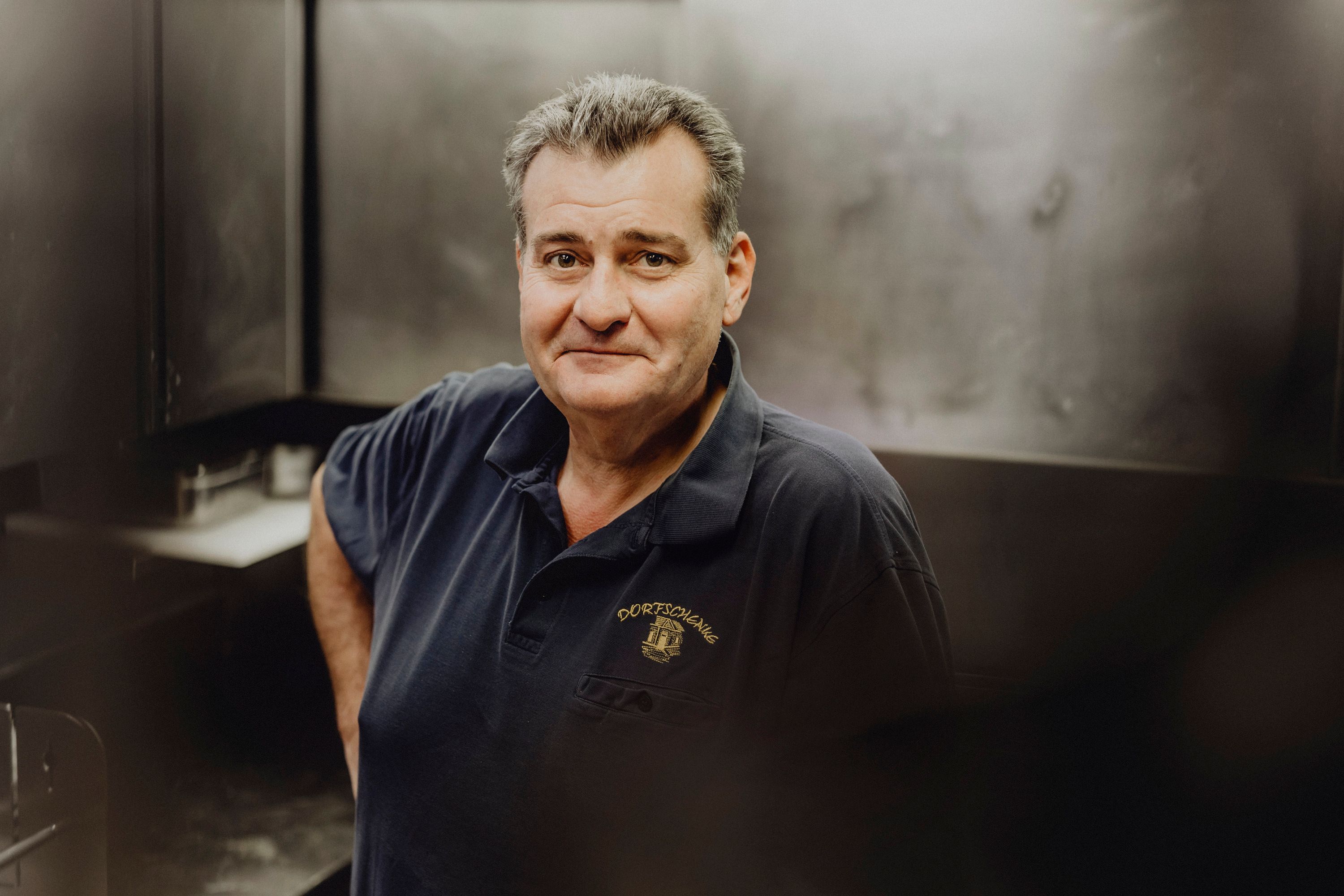 Man in a dark polo shirt with the "Dorfschenke" logo in a kitchen.