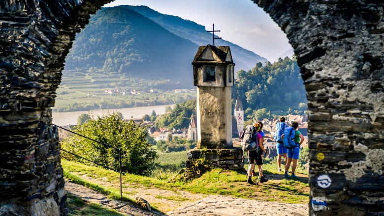 View through a stone gate onto a landscape with hills, a river and hikers.