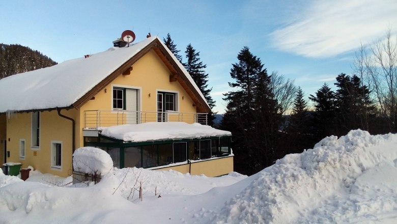 A yellow house with a snow-covered roof and balcony, surrounded by trees and snow.