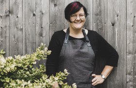 A woman with glasses and an apron stands smiling in front of a wooden wall next to a bush.