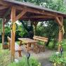 Wooden arbor in the garden with table and benches, surrounded by plants.