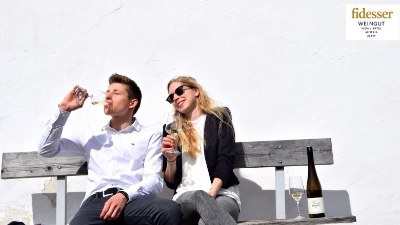 Two people are sitting on a bench drinking wine in front of a white wall.