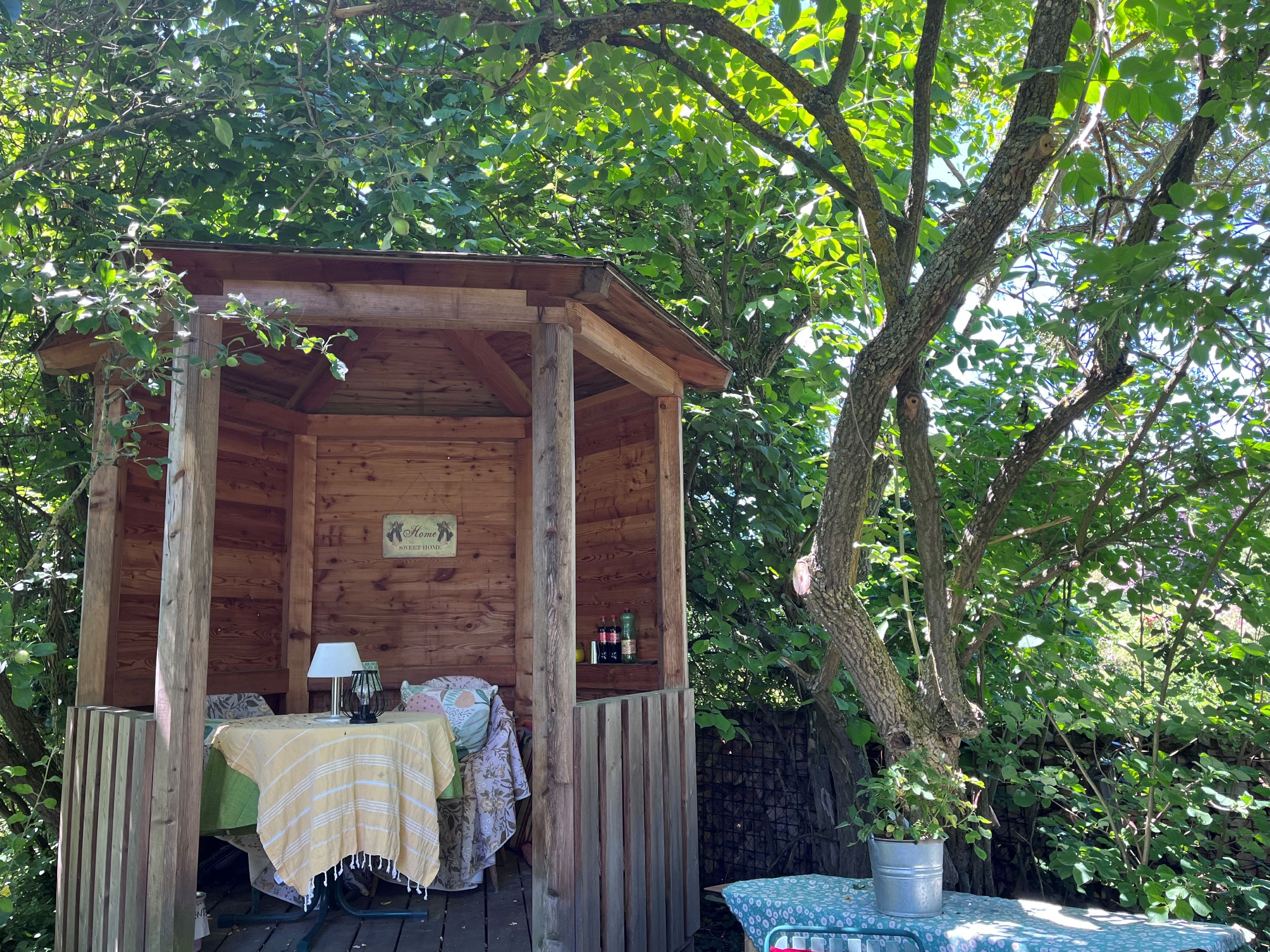 A cozy wooden pavilion in the garden, surrounded by trees and plants, with a table and chairs underneath.