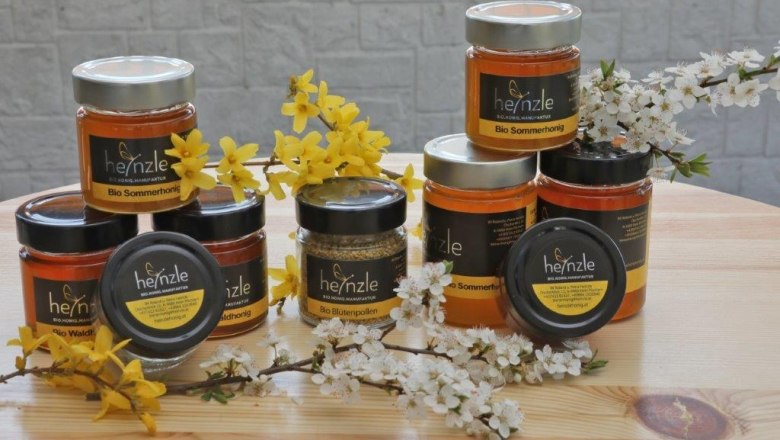 Various jars of honey from the Heinzle apiary on a wooden table, surrounded by yellow and white flowers.