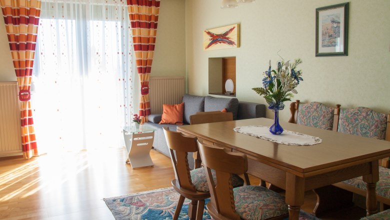 Living room, © Fam. Sulzbacher A cozy living room with wooden table, chairs, sofa and colorful curtains.