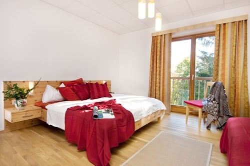 Bright bedroom with double bed, red pillows and blanket, wooden floor and large window with curtains.