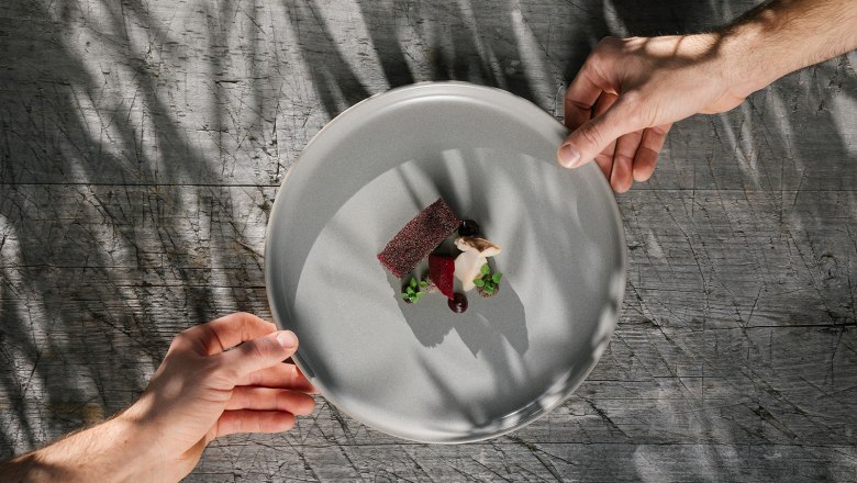 Two hands hold a plate with an artfully arranged gourmet dish above a wooden table.