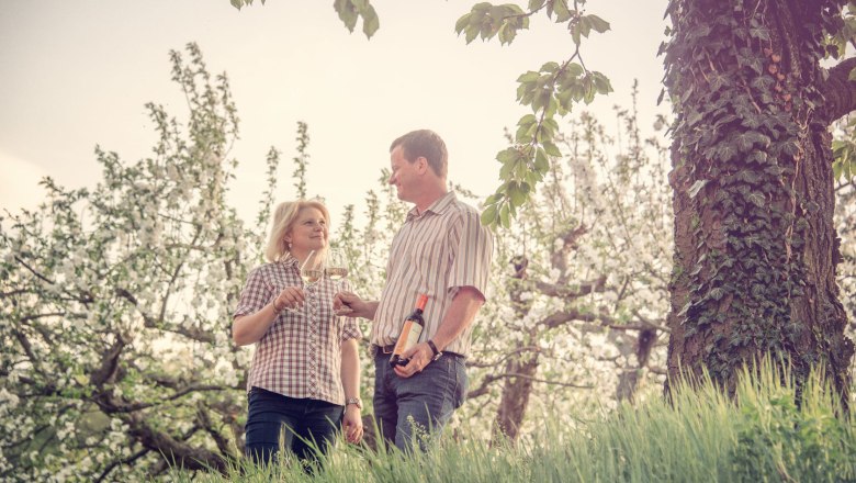 A couple stand in a blossoming orchard and clink glasses of wine while the man holds a bottle of wine.