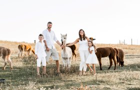 A family in white clothing stands in a meadow with alpacas.