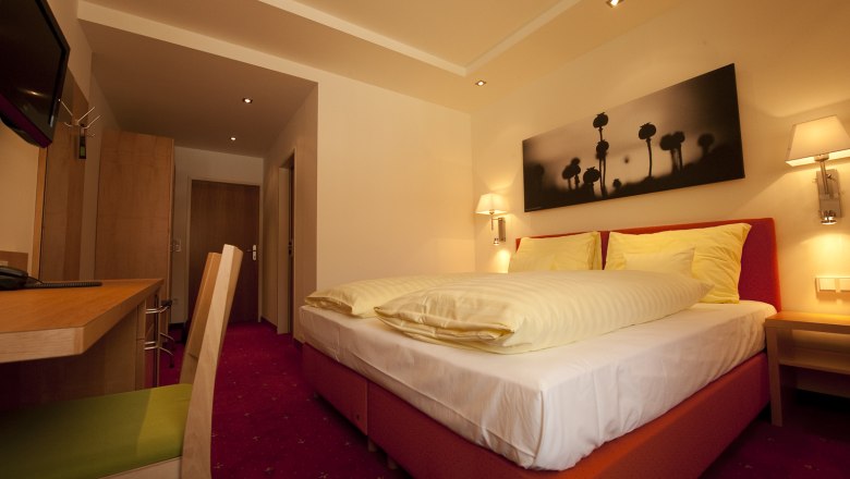 A cozy hotel room with a double bed, desk and mural. Warm lighting and red carpets create an inviting atmosphere.
