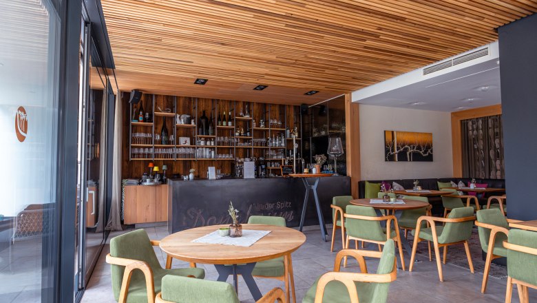 Interior view of the wine lounge with wooden elements, green chairs and a bar in the background.