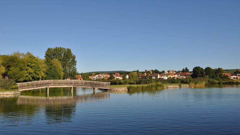 Wooden bridge over a quiet pond with a village in the background.