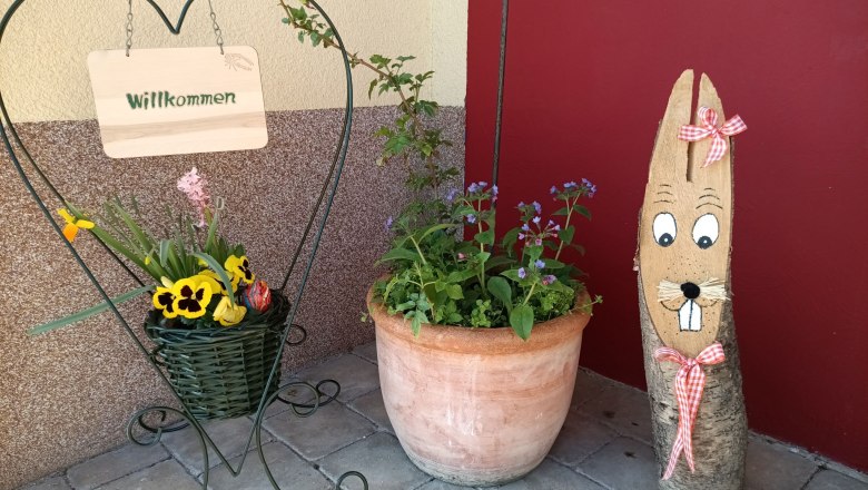 Welcome sign in the shape of a heart with flowers and wooden figure in front of a red wall.