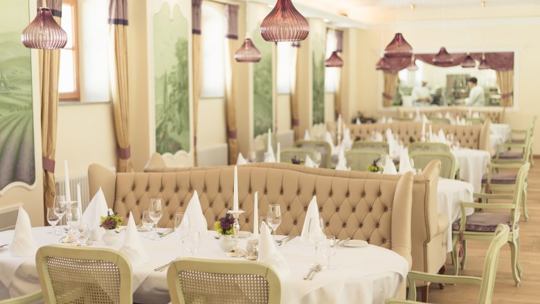 Elegant restaurant with laid tables, white tablecloths and candlesticks, beige upholstered benches and green chairs, decorative murals.