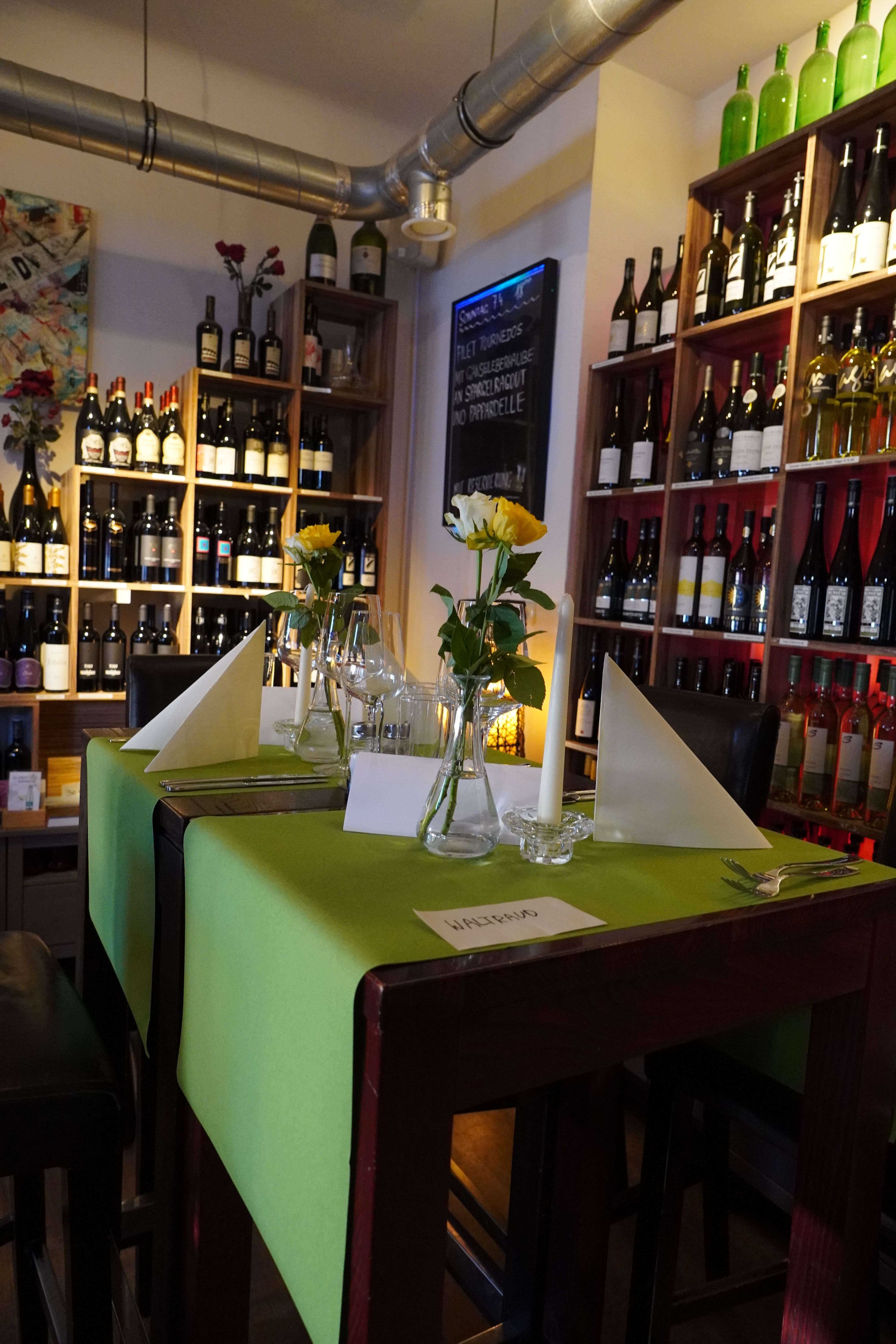A stylishly laid table in a café with a green tablecloth, yellow roses and bottles of wine in the background.