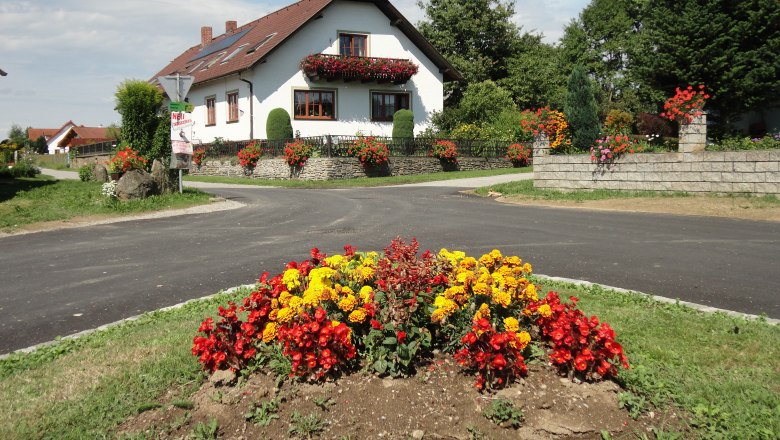 Guesthouse Reuberger, © Gästehaus Reuberger A white house with red roof tiles and floral decorations, surrounded by colorful flowerbeds and a green landscape.