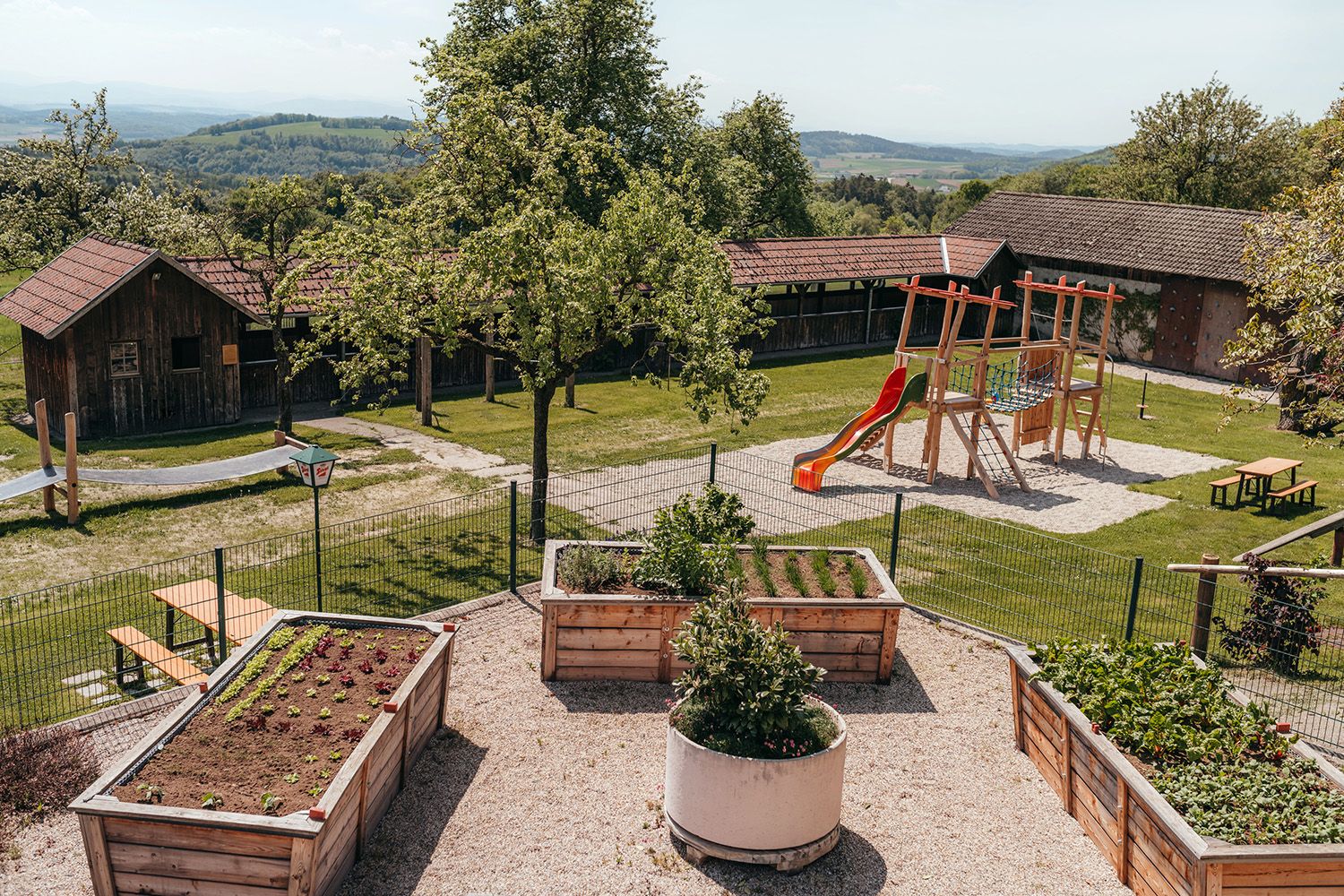 A playground with a slide and climbing frame next to raised beds in a garden with wooden huts in the background.