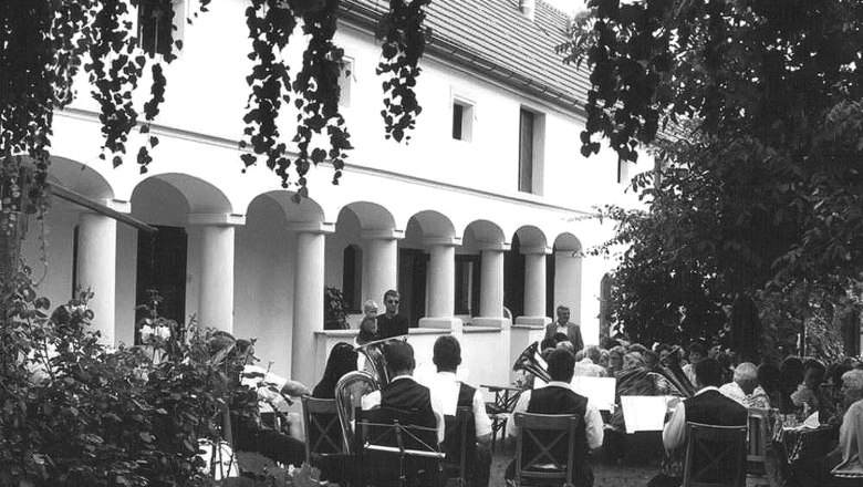 A group of people sit outside in front of a building with pillars while a brass band plays.