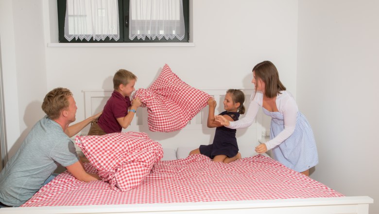 Family having pillow fight on a bed with red and white checkered bedding.