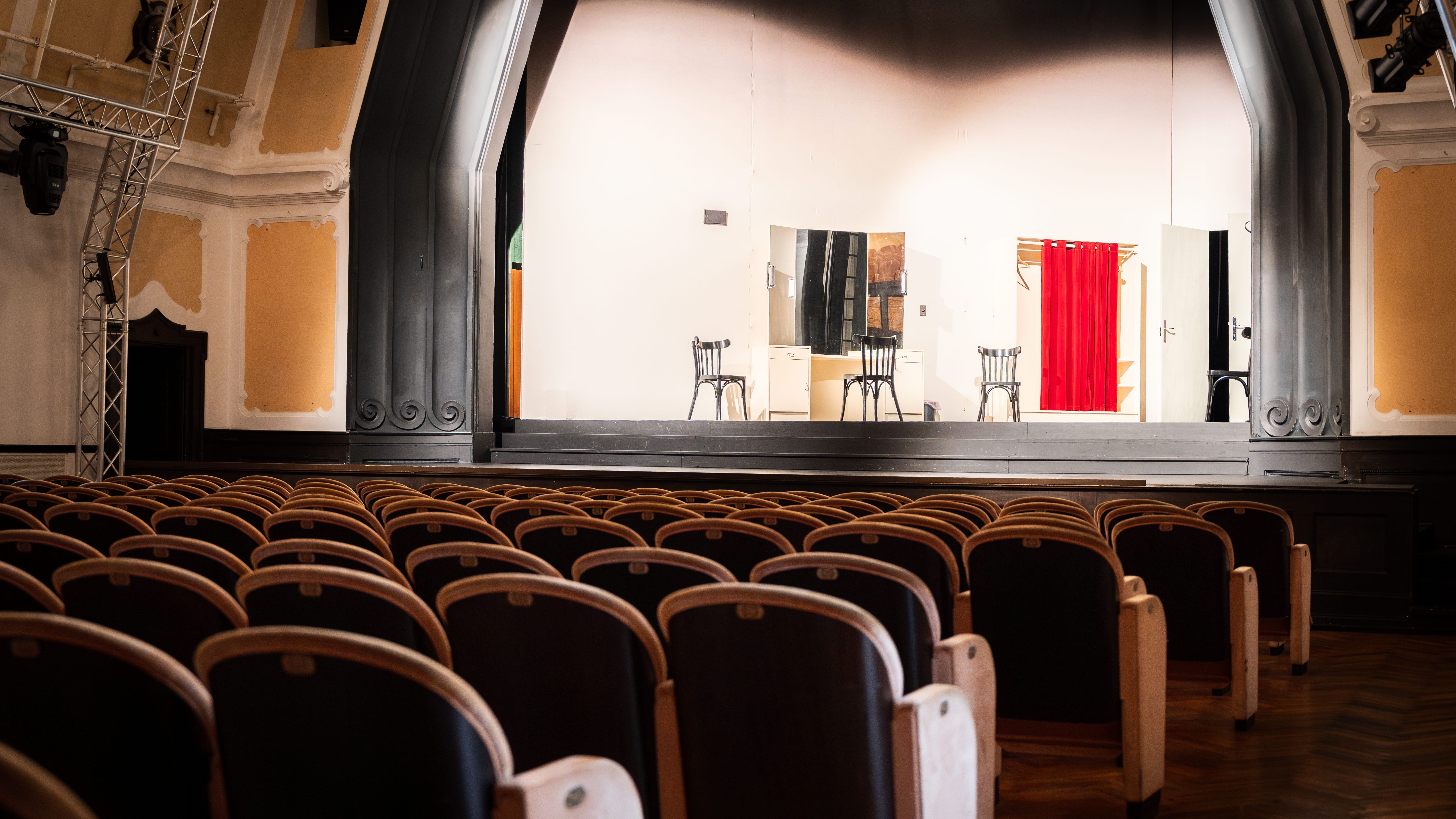 Empty theater hall with a stage on which two chairs and a red curtain can be seen.