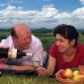 A couple having a picnic in a meadow with wine and a fruit basket, in the background a hilly landscape and a bicycle.