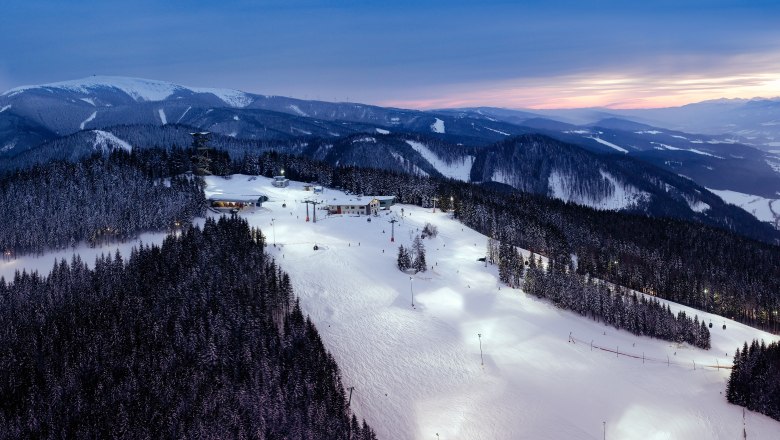 Illuminated ski slope at Semmering at dusk with snow-covered trees and mountains in the background.