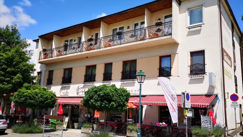Exterior view of a café with balcony and awning, surrounded by trees and street signs.