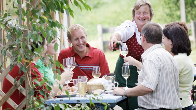 A group of people are sitting at an outdoor table enjoying wine. A woman pours wine and everyone smiles.