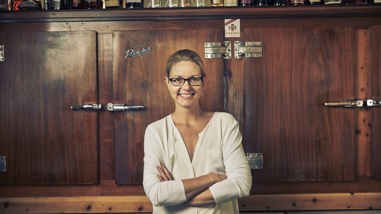 A woman with glasses stands smiling in front of a wooden cabinet with bottles on it.
