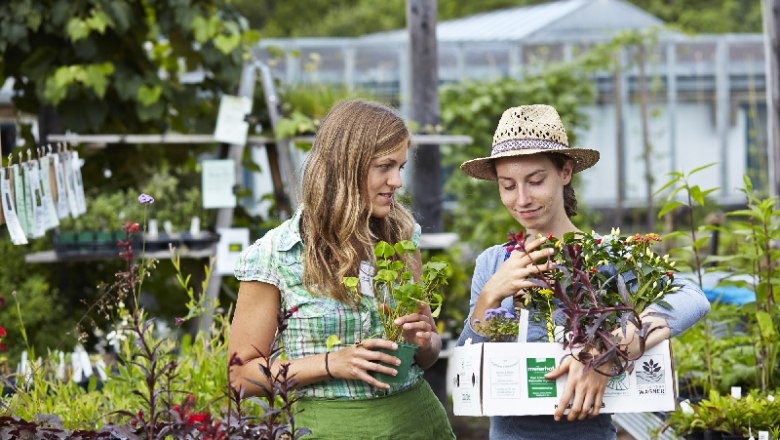 Two women in a garden store, one with a straw hat, hold plants in their hands.