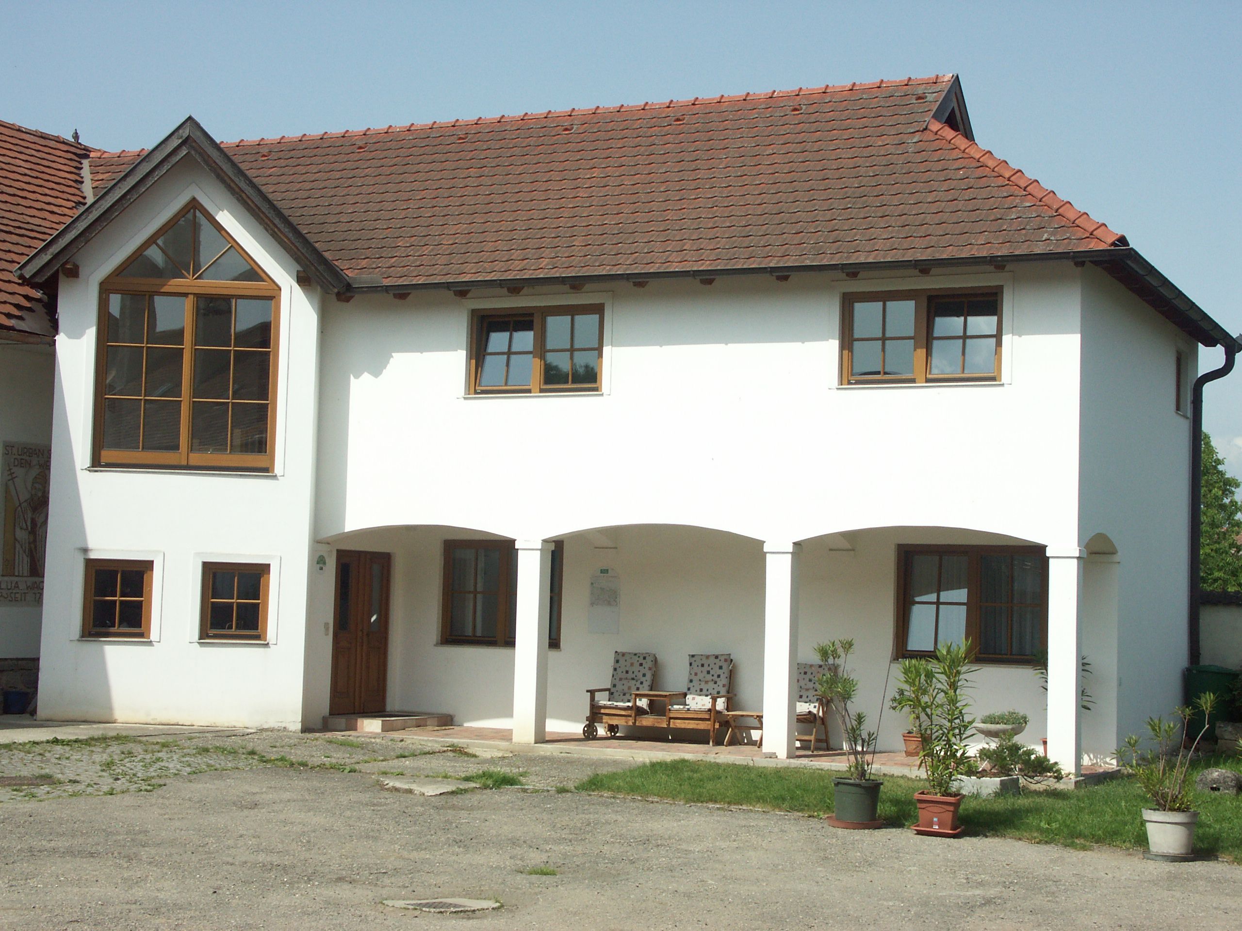A two-storey white building with red roof tiles and large windows. There are plants in pots in front of the house.