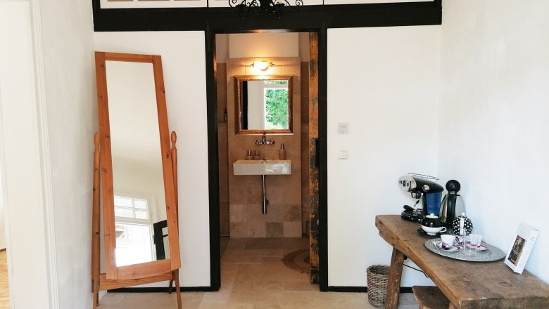 An anteroom with a large mirror, a coffee machine on a wooden table and a washbasin in the background.