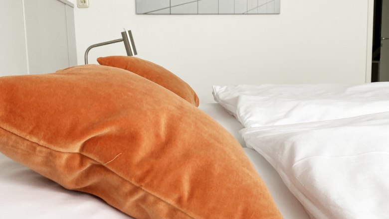 A bed with orange pillows and white bed linen in a guest room. A picture with hooks on tiles hangs on the wall.