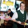 A waiter in a green vest serves a dish in a restaurant. Two men are sitting at a table in the background.