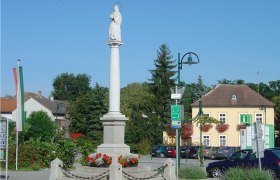 Statue on a column in a square in Haugsdorf, surrounded by buildings and trees.