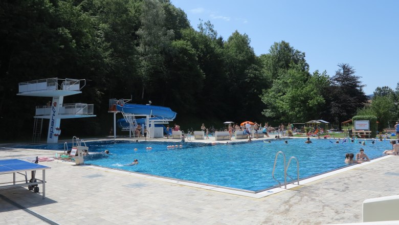 An outdoor pool with diving platform, slide and lots of bathers on a sunny day.