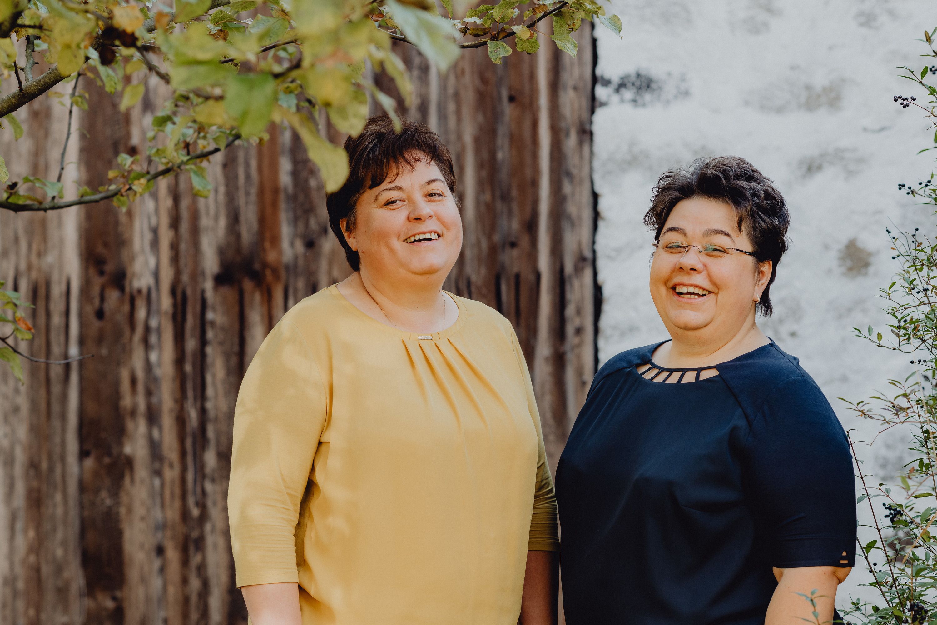 Two smiling women stand in front of a wooden wall outside.