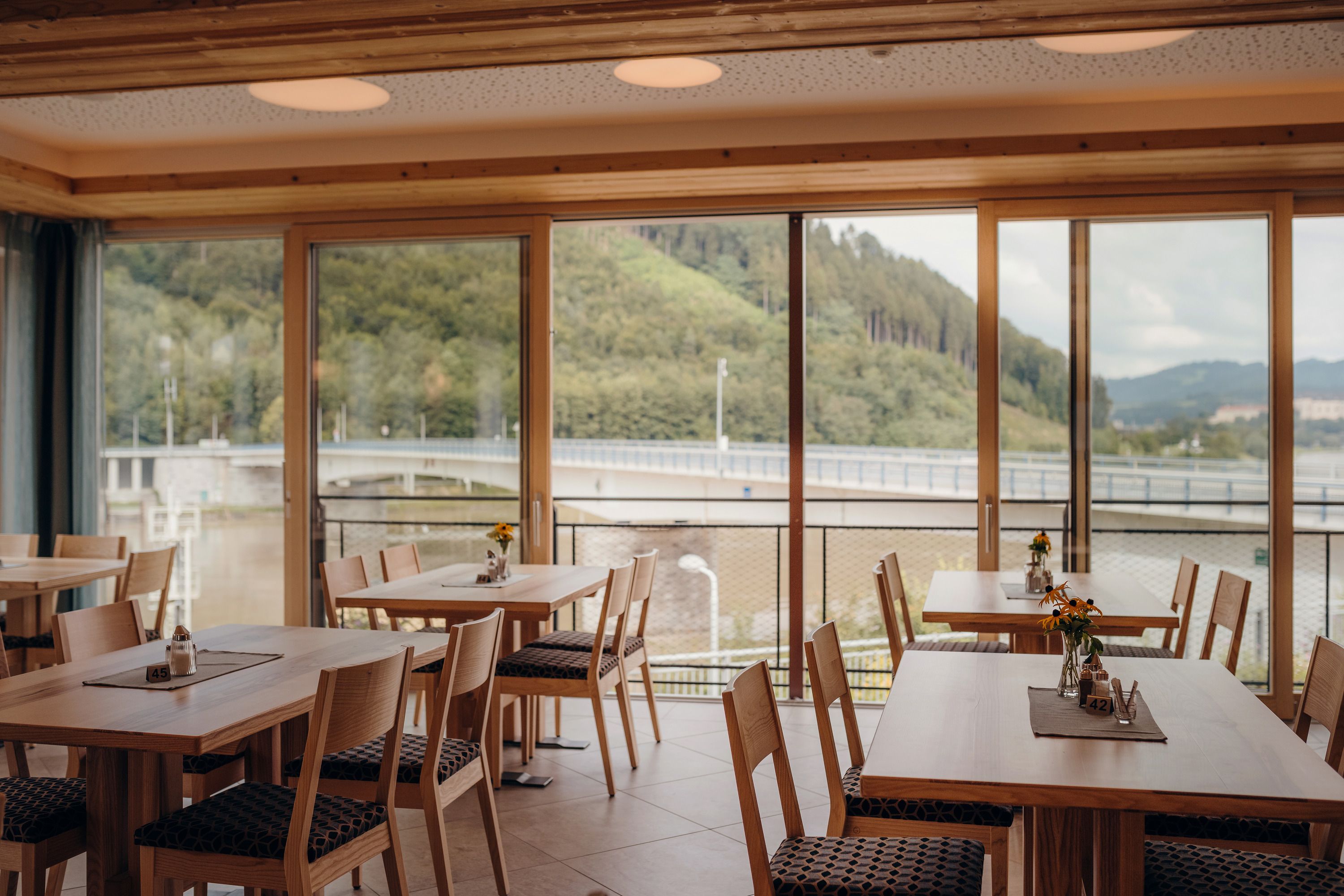 Interior view of an inn with large windows and a view of a bridge and wooded hills.