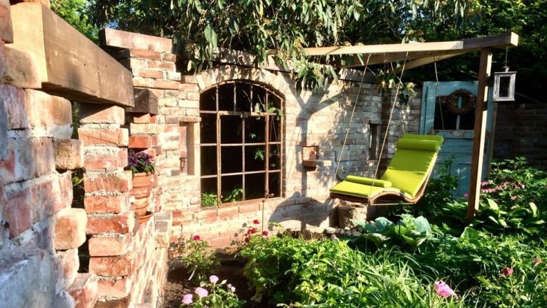 A cozy garden with an old brick wall, a window arch and a green lounger under a wooden frame.