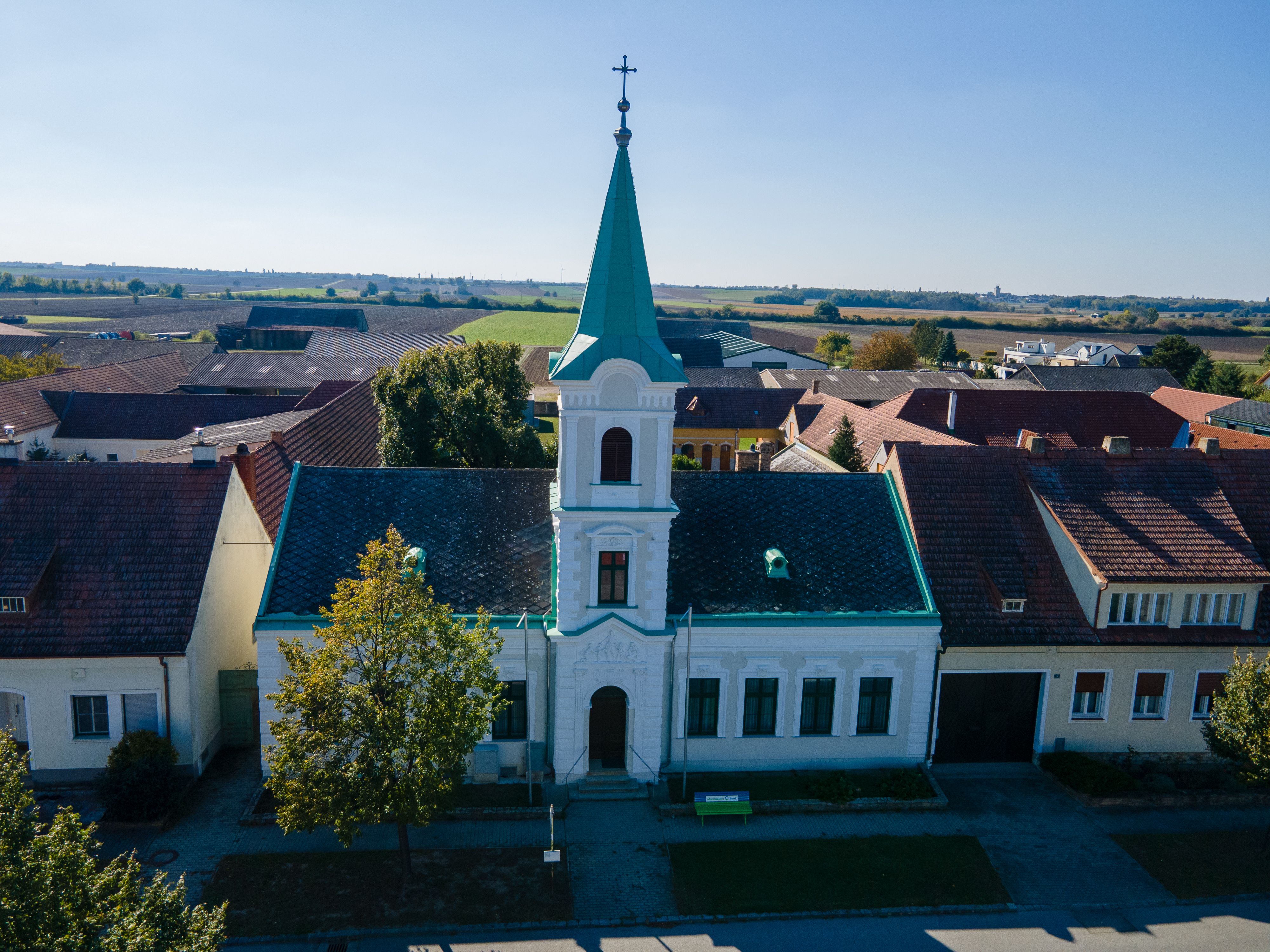 A church in the town of Weiden an der March with a green tower and a cross, surrounded by houses and fields.