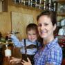 A woman holds a child in lederhosen by a beer tap. Bottles of spirits can be seen in the background.