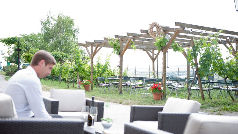 A man sits in a garden with vines and empty tables in the background.