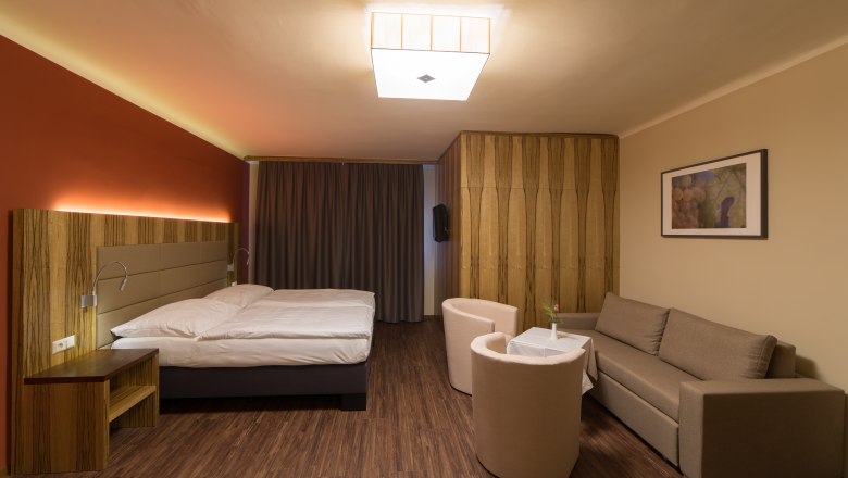 Modern furnished hotel room with double bed, sofa, two armchairs and a small table. Wooden floor and warm lighting.
