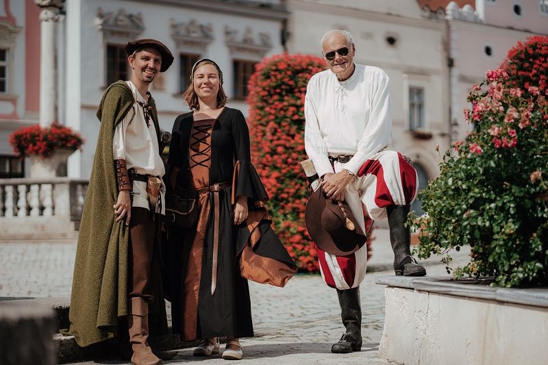 Three people in historical costumes stand in front of a square decorated with flowers.
