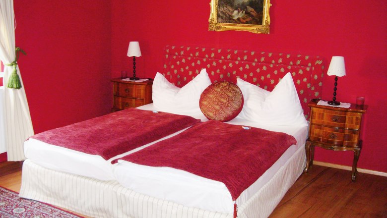 A bedroom with red walls, a double bed with red blankets and a painting on the wall.