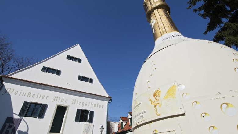 Large champagne bottle in front of a white building with the inscription 'Weinkeller Max Riegelhofer'.