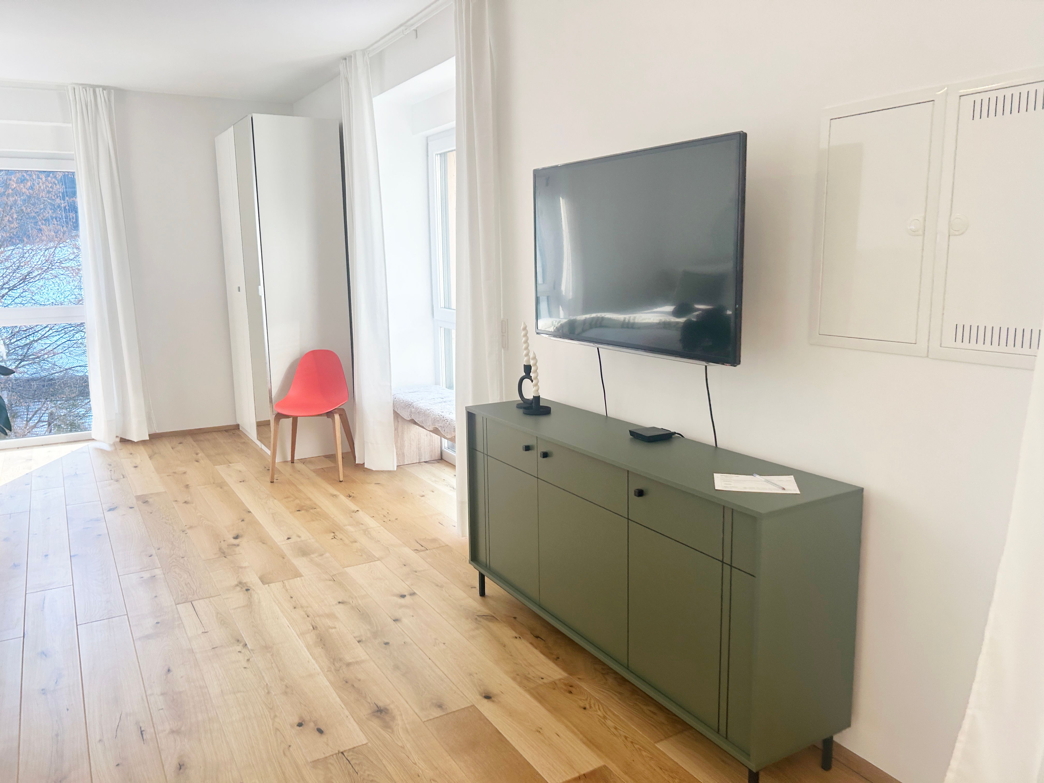 Modern living room with wooden floor, green sideboard, wall-mounted TV and red chair in front of a mirror.