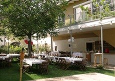 A guest garden with covered tables and chairs under a tree, next to a building with a balcony.