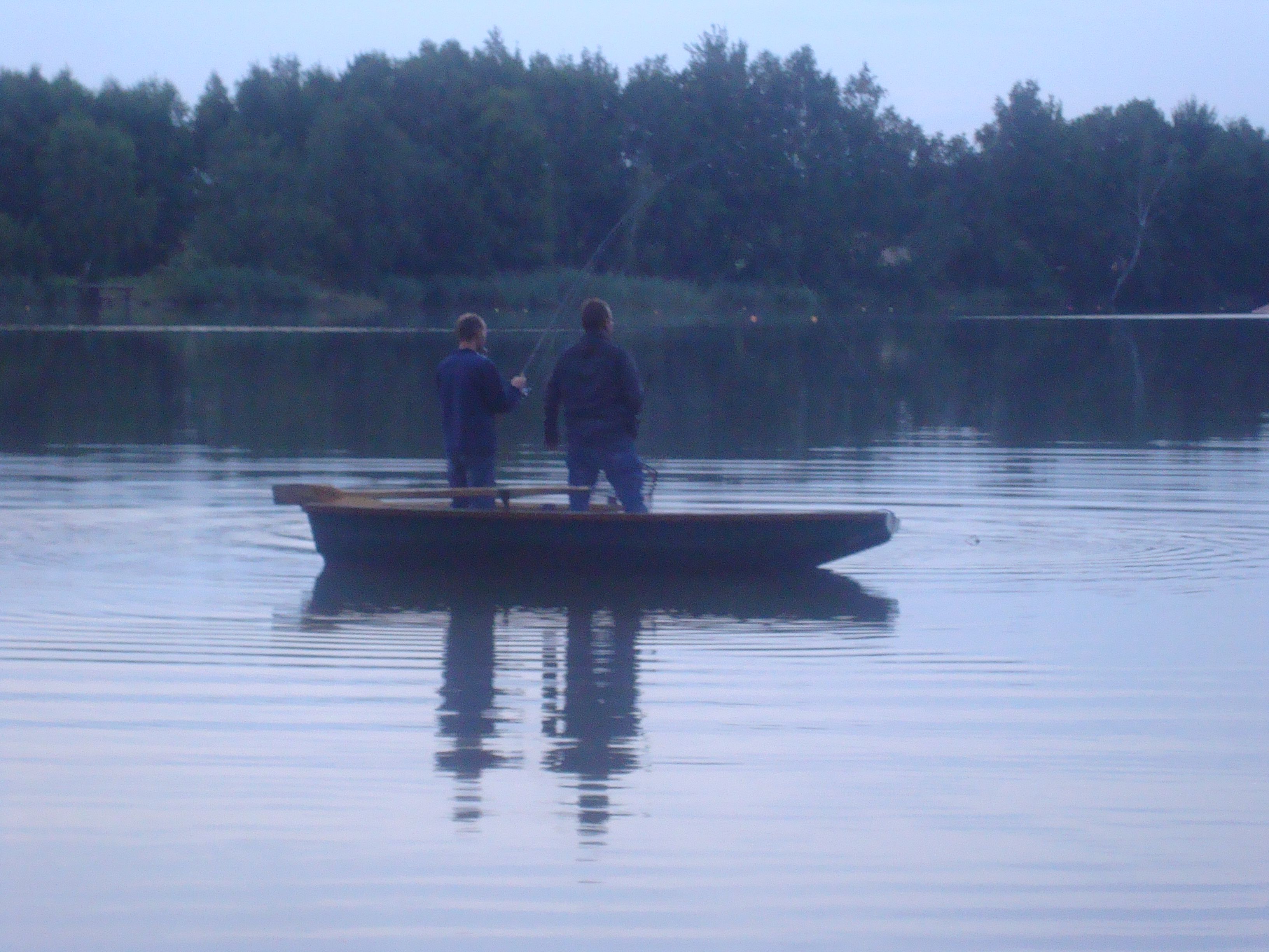 Two people fishing on a boat on a calm lake surrounded by trees.
